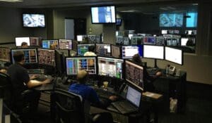 This image portrays an IT department. There are a few people working on laptops and each of them has two to three additional monitors in their workspace.